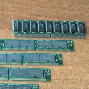 May include: Five green computer memory modules, or RAM sticks, are arranged in a row on a wooden surface. The modules are all the same size and shape, and they have black chips on them. The modules are arranged in a descending order of size, with the largest module at the top of the image and the smallest module at the bottom.