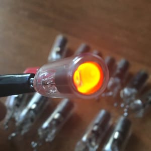May include: A close-up of a small, clear glass bulb with a glowing orange filament inside. The bulb is connected to a black wire and is surrounded by other similar bulbs.