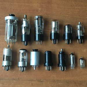 Op de afbeelding: Een verzameling van 13 vintage vacuümbuizen, sommige met glazen en metalen behuizingen, andere met zwarte kunststof behuizingen. De buizen zijn in twee rijen op een houten oppervlak gerangschikt.