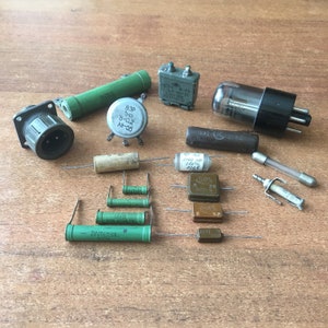 Puede incluir: Surtido de componentes electrónicos, que incluyen resistencias, condensadores, transistores y un tubo de vacío. Los componentes están dispuestos sobre una superficie de madera. Algunos de los componentes tienen texto impreso, como "B3P 1M0 B-0.2 xil-55", "2700 20 240% 6006" y "2W1C7591".