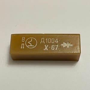 May include: An amber-coloured rectangular electronic component. It features printed text and symbols, including "Д1004" and "X-67", plus a diode symbol. The component is likely used in electronics.