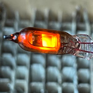 May include: A glowing orange and yellow vacuum tube with a clear glass exterior. The tube has a pointed end and a clear, bulbous end with red wires visible inside. The light source is a bright, rectangular shape.