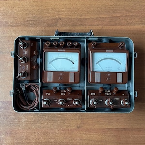 Puede incluir: Equipo de prueba eléctrica vintage marrón y plateado en una caja metálica. La caja contiene múltiples medidores, diales e interruptores. Los medidores tienen caras blancas con marcas y agujas negras. El equipo parece estar en buenas condiciones.