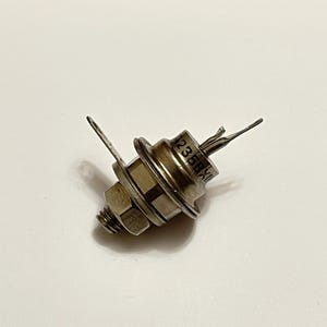 以下が含まれることがあります： ナットとボルトのベース、円形の本体、2つの平らな金属タブが外側に伸びた金属製の電子部品。コンポーネントの表面には「1235BX」の文字が印刷されています。このオブジェクトは、おそらく電気回路で使用されます。