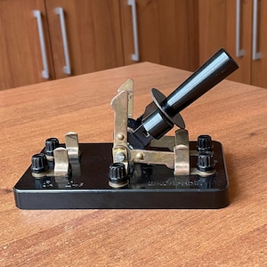 May include: A vintage black telegraph key with brass components and a black handle. The key is mounted on a rectangular black base with several terminals. The text "ШКОЛЬНЫЙ" is visible on the base.
