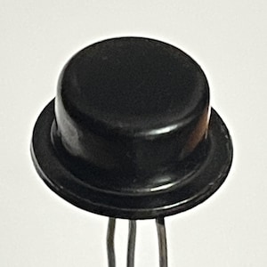 Puede incluir: Un componente electrónico negro con una parte superior redondeada y una base circular. Tres pines plateados se extienden desde la parte inferior. El componente está sobre un fondo blanco liso.