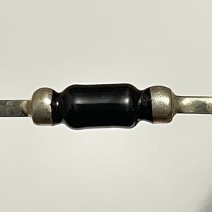 May include: Close-up of a black electronic component with silver-colored terminals. The component has a cylindrical black body with silver-colored ends. The terminals extend from each end of the component.