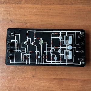Op de afbeelding: Een zwarte printplaat met verschillende elektronische componenten, waaronder weerstanden, condensatoren en transistoren. De printplaat heeft een zwarte achtergrond met witte lijnen en markeringen. Er zijn verschillende zwarte en witte draden aangesloten op de printplaat.
