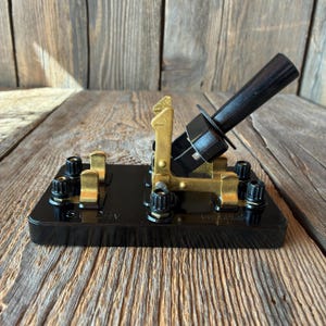 Pode incluir: Um interruptor de faca elétrico preto montado numa base retangular preta com pontos de contacto em latão e uma pega preta. A base apresenta o texto em relevo 5A 24V. O mecanismo inclui uma alavanca de latão e um punho de plástico preto.