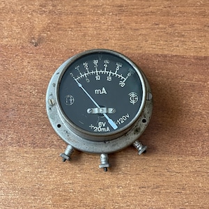 Può includere: Un vecchio misuratore di mA analogico con un quadrante grigio scuro e un ago che punta al segno di 5 mA. Il misuratore ha un design circolare con un involucro in metallo e tre terminali. Il quadrante mostra il testo "mA", "6V" e "+20mA".