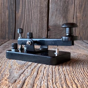 May include: A black telegraph key mounted on a rectangular base, featuring a prominent knob at one end, a central metal pivot mechanism, and two adjustable screw terminals with knurled caps for electrical connections, all finished in a glossy black.