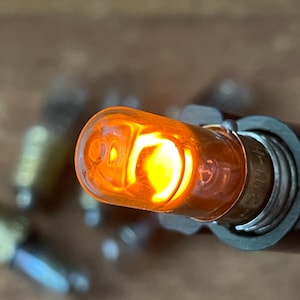 May include: Close-up of a lit amber-colored automotive light bulb. The bulb is encased in a clear, rounded glass and emits a bright, warm glow. The base of the bulb is silver and attached to a metal housing.