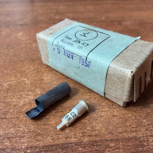 Könnte beinhalten: Eine rechteckige braune Pappschachtel mit einem hellblauen Papieretikett, das den Text "Тип ДК-С2", "Колич. 10" und "9 НОЯ 1958" trägt. Neben der Schachtel liegen ein kleiner dunkelgrauer Metallzylinder und ein silberfarbenes Metallbauteil mit der Aufschrift "C2" und "58".