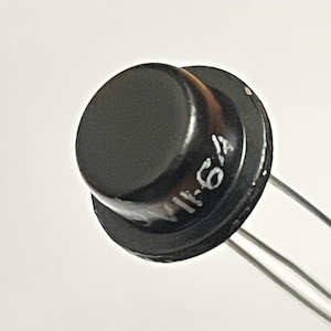 Könnte beinhalten: Nahaufnahme eines schwarzen elektronischen Bauteils mit dem weißen Text "11-64". Das Bauteil hat eine kreisförmige Oberseite und Metallanschlüsse. Der Hintergrund ist einfarbig hell.