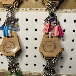 May include: Keychains with colorful tassels and gemstone beads. Each keychain has a silver ring, a small tassel, and a cluster of gemstones. The tassels come in various colors, including blue, pink, green, white, and black.