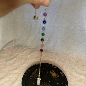 May include: A silver pendulum with a chain of colorful beads, including purple, blue, green, and red, is suspended above a black divination board. The board features gold celestial designs and the words "YES", "NO", and "TRY AGAIN".