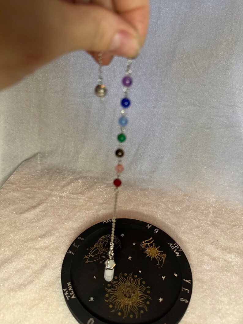 May include: A silver pendulum with a chain of colorful beads, including purple, blue, green, and red, is suspended above a black divination board. The board features gold celestial designs and the words "YES", "NO", and "TRY AGAIN".