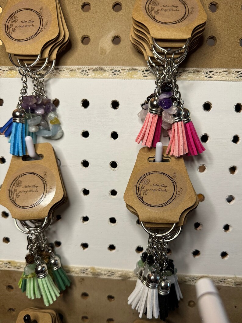 May include: Keychains with colorful tassels and gemstone beads. Each keychain has a silver ring, a small tassel, and a cluster of gemstones. The tassels come in various colors, including blue, pink, green, white, and black.