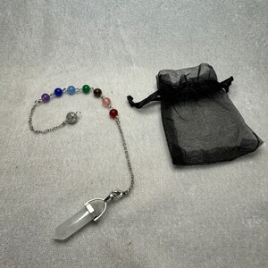 May include: A pendulum with a clear crystal point and a chain adorned with colorful beads. The chain is connected to a series of small, round, colored beads, representing the chakras. A black drawstring bag is next to the pendulum.