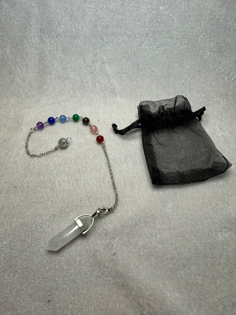 May include: A pendulum with a clear crystal point and a chain adorned with colorful beads. The chain is connected to a series of small, round, colored beads, representing the chakras. A black drawstring bag is next to the pendulum.
