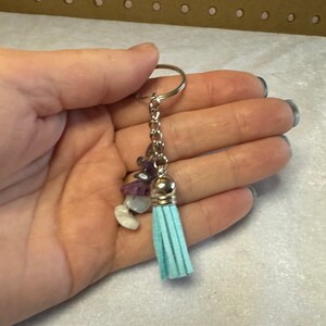May include: A silver keychain with a light blue tassel and a cluster of small, colorful gemstones. The gemstones include purple, white, and clear stones. The keychain is held in a hand.