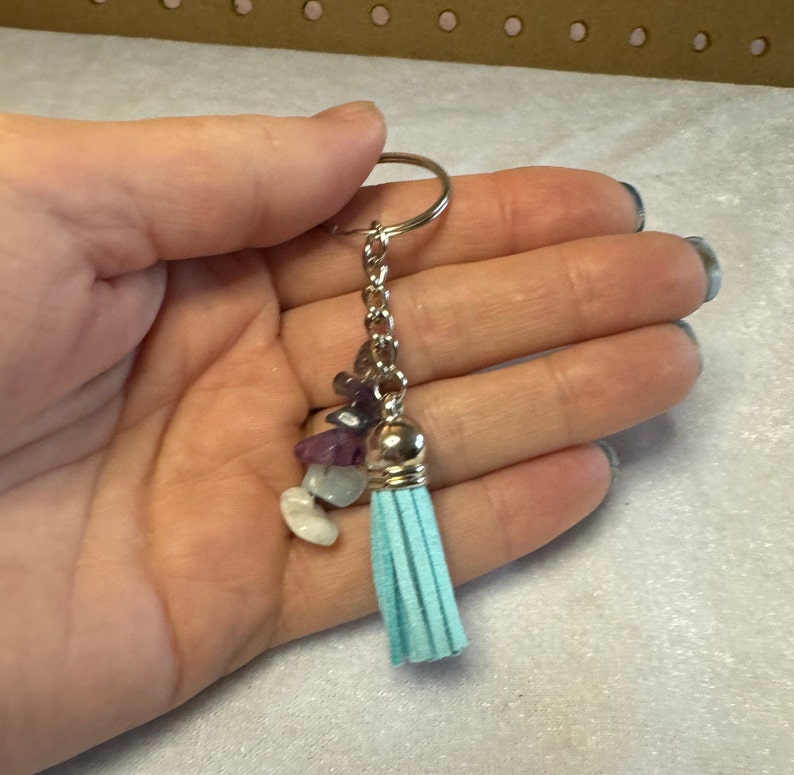 May include: A silver keychain with a light blue tassel and a cluster of small, colorful gemstones. The gemstones include purple, white, and clear stones. The keychain is held in a hand.