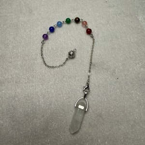 May include: A divination pendulum featuring a clear quartz crystal point suspended by a silver chain. The chain is adorned with a series of coloured beads. The pendulum is designed for spiritual practices.