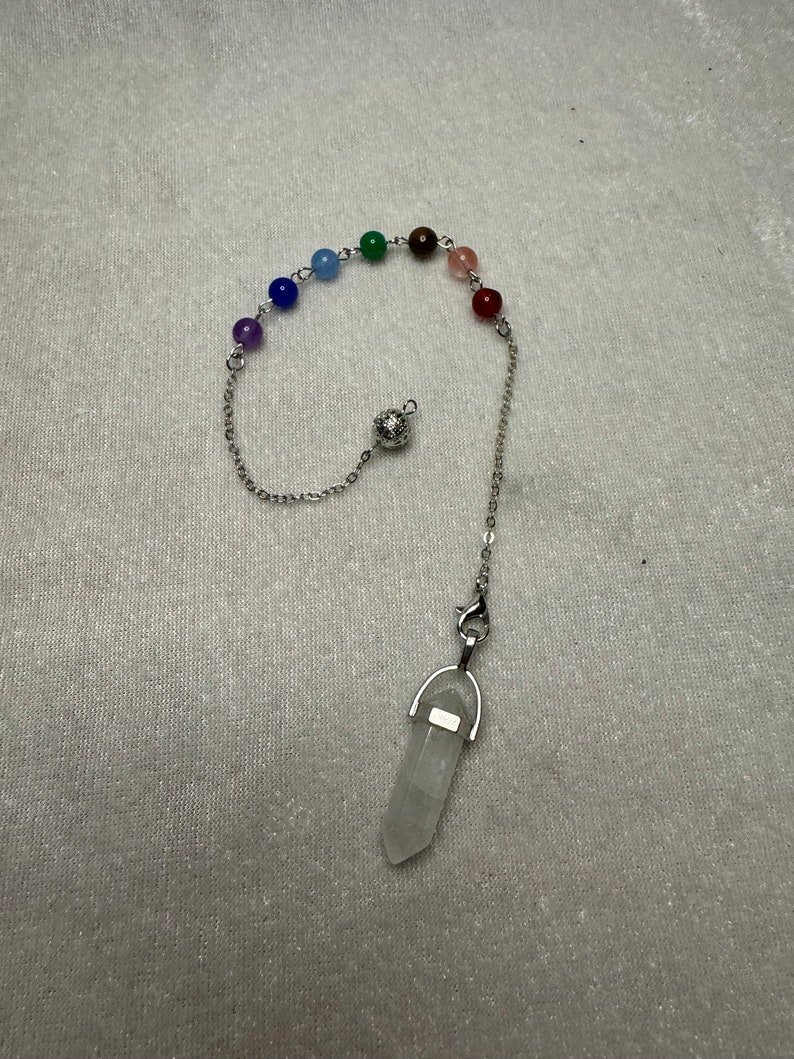 May include: A pendulum with a clear quartz crystal point and a chain with colorful beads. The beads are arranged in a rainbow spectrum. The pendulum is designed for divination and spiritual practices.