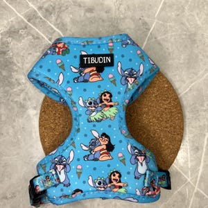 Op de afbeelding: Een lichtblauw huisdierentuigje met een herhalend patroon van Stitch- en Lilo-figuren, ijshoorntjes en kleine stippen. Het tuigje is voorzien van een zwart rechthoekig label met de witte tekst TIBUDIN en zwarte plastic gespen aan de zijkanten.