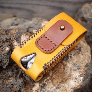 Op de afbeelding: Een gele leren multitool-schede met een bruine leren flap die is vastgezet met een messing knop. De schede heeft een gestikte rand en een multitool is gedeeltelijk zichtbaar. Het item ligt op een ruwe, grijze rots.