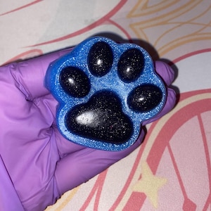 Könnte beinhalten: Eine blaue Pfotenabdruck-förmige Seife mit schwarzen Glitzer, die in die Seife eingebettet sind. Die Pfote hat fünf Zehen.