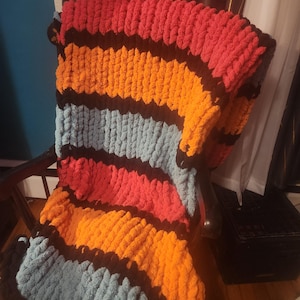Può includere: Una coperta a maglia grossa a righe rosse, arancioni, azzurre e nere, drappeggiata su una sedia. La lana spessa della coperta le conferisce un aspetto strutturato e accogliente. Ideale per aggiungere calore e stile a qualsiasi spazio.