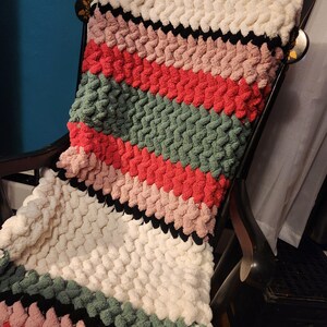 Könnte beinhalten: Eine grob gestrickte Decke auf einem Schaukelstuhl. Die Decke hat horizontale Streifen in Weiß, Schwarz, Rosa, Rot und Grün. Die weiche, strukturierte Decke ist über den Stuhl drapiert und zeigt ihr gemütliches Design und ihre Farbpalette.
