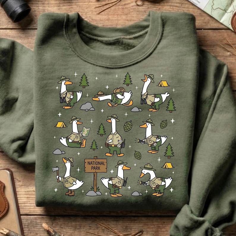 Op de afbeelding: Olijfgroene sweatshirt met een herhaald patroon van cartoon ganzen gekleed als parkwachters. De ganzen worden in verschillende poses afgebeeld, interactie met bomen, kaarten en kampeerspullen. Een bord leest "National Park".
