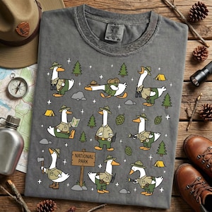 Op de afbeelding: Grijs t-shirt met een grillig ontwerp van ganzen gekleed als parkwachters. De ganzen worden afgebeeld in verschillende buitenactiviteiten, zoals kamperen en wandelen. Het shirt bevat een "National Park"-bord.