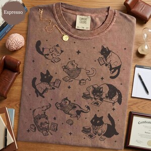 May include: A brown crew neck t-shirt featuring a black graphic print of multiple cats in various poses, some wearing sweaters or glasses and holding clipboards or books, interspersed with small star shapes. The shirt has a Comfort Colors label.