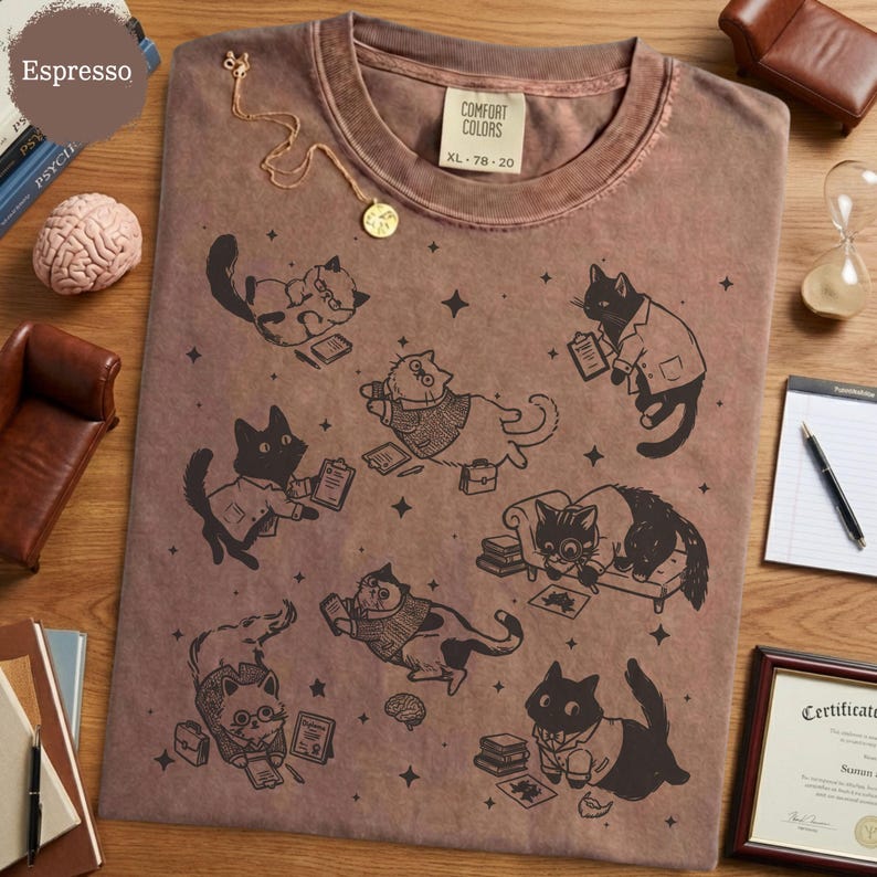 May include: A brown crew neck t-shirt featuring a black graphic print of multiple cats in various poses, some wearing sweaters or glasses and holding clipboards or books, interspersed with small star shapes. The shirt has a Comfort Colors label.