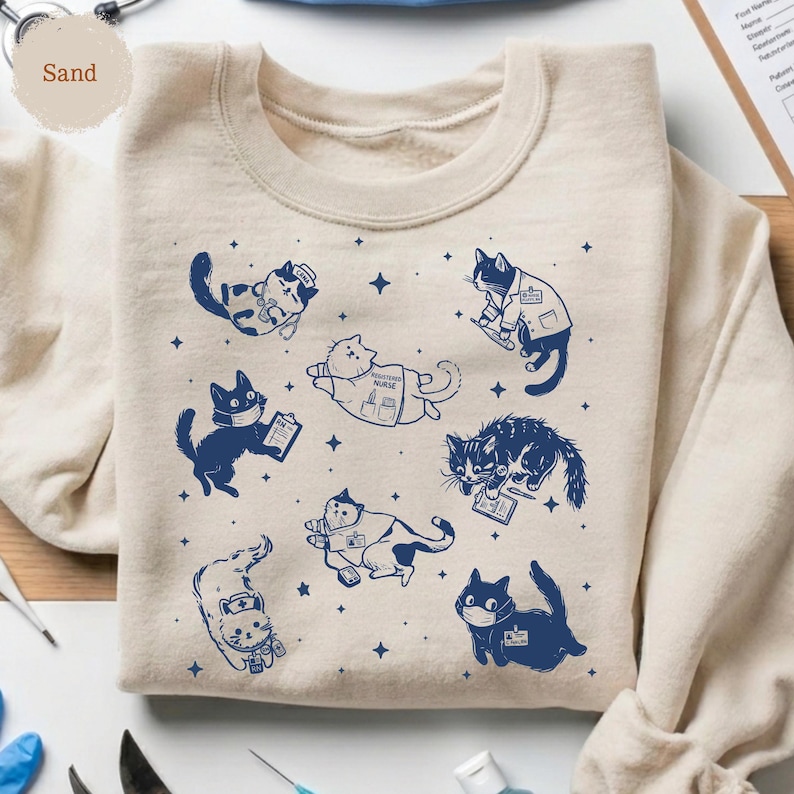 May include: Sand-colored sweatshirt featuring navy blue illustrations of cats in various medical roles, including nurses and doctors. The cats are depicted with stethoscopes, clipboards, and face masks, surrounded by star accents. The word "Sand" is in the upper left corner.
