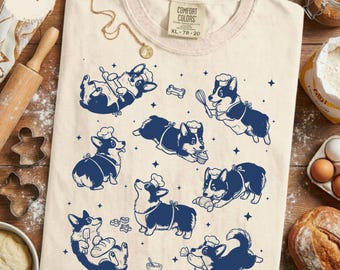 Könnte beinhalten: Ein elfenbeinfarbenes T-Shirt mit einem dunkelblauen Grafikdruck von mehreren Corgi-Hunden, die Kochmützen tragen und backen. Die Hunde sind in verschiedenen Posen dargestellt, unter anderem mit Nudelhölzern, Schneebesen und Backwaren, umgeben von kleinen Sternsymbolen.