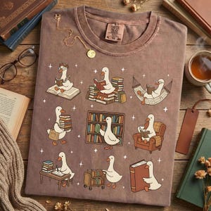 Puede incluir: Una camiseta marr&oacute;n con un dise&ntilde;o caprichoso de gansos leyendo libros en varios entornos. Los gansos se representan con gafas y leyendo en una hamaca, sobre montones de libros y en un escritorio. La camiseta tiene una etiqueta Comfort Colors.