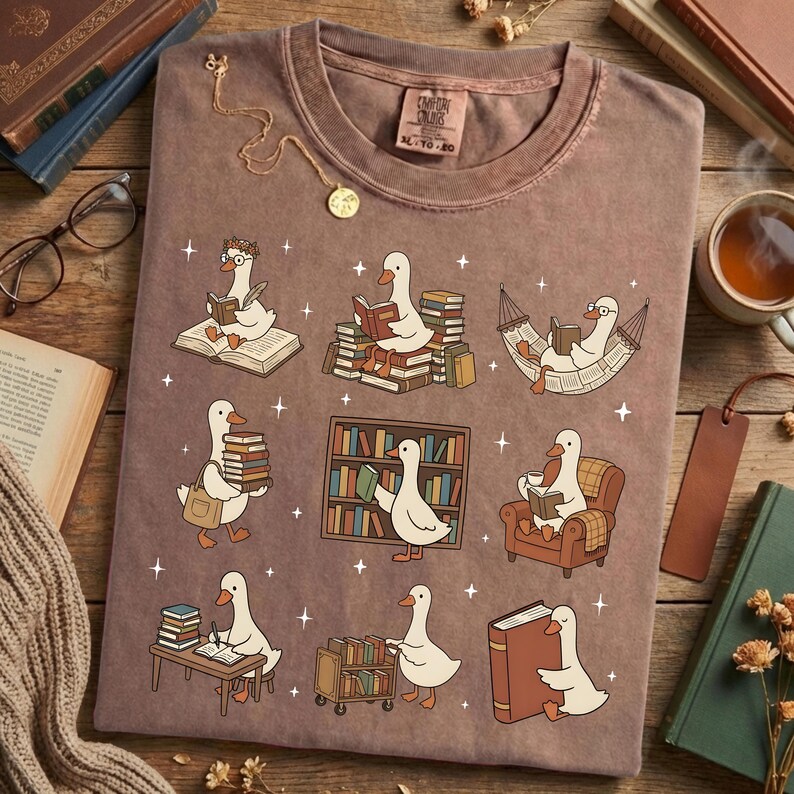 Puede incluir: Una camiseta marr&oacute;n con un dise&ntilde;o caprichoso de gansos leyendo libros en varios entornos. Los gansos se representan con gafas y leyendo en una hamaca, sobre montones de libros y en un escritorio. La camiseta tiene una etiqueta Comfort Colors.