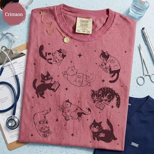 May include: Crimson-colored Comfort Colors t-shirt with a crew neck, featuring illustrations of cats in various medical roles. The shirt includes the text "Comfort Colors" and size XL 78-20. A gold necklace and medical tools are also visible.