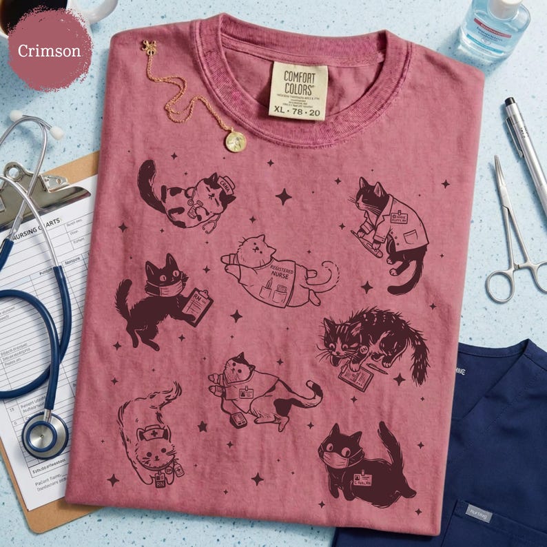 May include: Crimson-colored Comfort Colors t-shirt with a crew neck, featuring illustrations of cats in various medical roles. The shirt includes the text "Comfort Colors" and size XL 78-20. A gold necklace and medical tools are also visible.