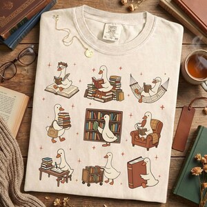 Puede incluir: Camiseta de color crema con un dise&ntilde;o caprichoso de gansos de dibujos animados leyendo libros en varios entornos. Los gansos est&aacute;n representados con gafas y accesorios, rodeados de libros y muebles. La camiseta tiene el texto "CAMPY COLORS" y la talla "XL 70-20".