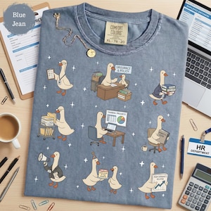 May include: A blue jean colored t-shirt featuring a graphic print of various cartoon geese performing office tasks. The geese are depicted with items like a grievance and policy sign, a training progress chart, a benefits guide, and an employee of the month cake.