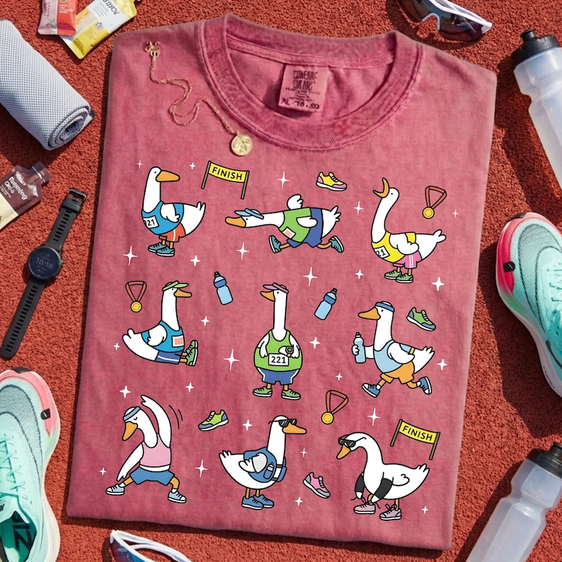 May include: A maroon t-shirt featuring cartoon geese in athletic gear, engaged in various running and exercise activities. The geese are depicted with medals, water bottles, and running shoes. The shirt also has the text "CONFORT COLORS" and size information.