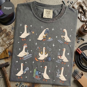May include: A charcoal gray crew neck t-shirt featuring a graphic print of nine white geese performing various welding and metalworking tasks, including using torches, hammers, and grinders, surrounded by small white star icons.