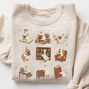 Puede incluir: Sudadera beige con un dise&ntilde;o caprichoso de gansos de dibujos animados leyendo libros en varios escenarios. Los gansos se representan con gafas, en una hamaca y cerca de estanter&iacute;as. El dise&ntilde;o incluye peque&ntilde;os acentos de estrellas.