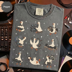 May include: Gray Comfort Colors t-shirt featuring a whimsical design of geese as DJs. The geese are depicted with headphones, turntables, and boomboxes. The shirt has a vintage, worn look, with the size XL-78-20 printed on the tag.