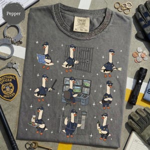 May include: Pepper-colored Comfort Colors t-shirt featuring cartoon geese dressed as correctional officers in various poses. The geese are depicted with hats, uniforms, and accessories like a baton, coffee, and a radio. The shirt has a distressed look.